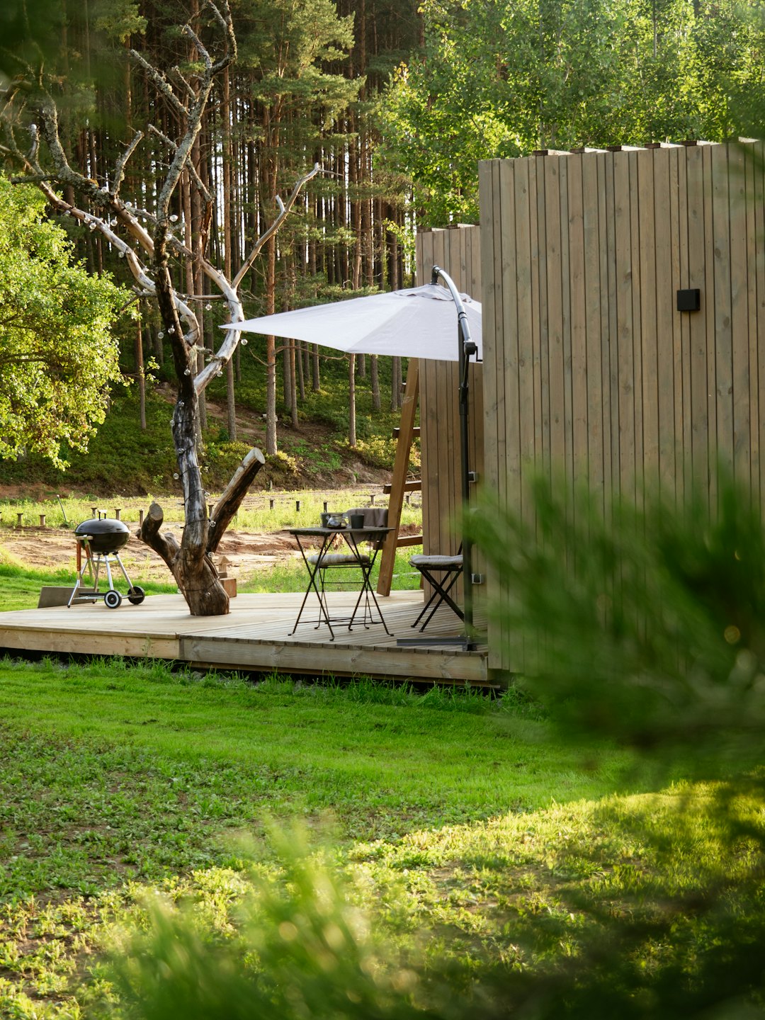 Außendeck mit Grill, Sonnenschirm und Bistrotischgarnitur in der Nähe eines Baumes.